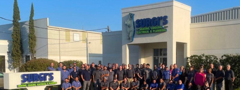 Large group of employees standing in front of Surgi's business location with signage for heating, cooling, electrical and plumbing services.
