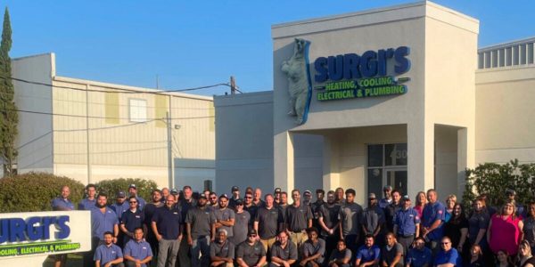 Large group of employees standing in front of Surgi's business location with signage for heating, cooling, electrical and plumbing services.